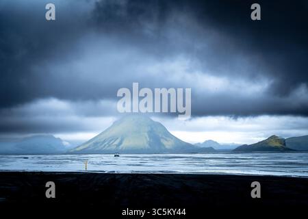 View of the mysterious green mountain shrouded in mist, standing guard over the dark, still waters under a stormy sky, Kirkjubaejarklaustur, Skaftárhreppur, Iceland. Stock Photo