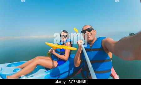 A man and woman are kayaking on a lake, wearing life jackets and holding paddles. The man is taking a selfie with his phone Stock Photo