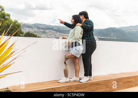 Pregnant woman with a prosthetic leg points at the cityscape while her partner embraces her, enjoying a scenic viewpoint Stock Photo