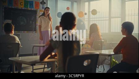 Female Teacher Finishing the Lesson, Announcing the End of School Day to Diverse Kids. Elementary School Students Standing Up, Packing Their Backpacks and Leaving Modern Classroom. Slow Motion. Stock Photo