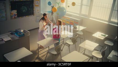 End of the School Day: Elementary School Girl Packing Her Backpack After Lesson and Leaving Modern Classroom. Female Teacher Helping the Girl to Pack Her School Supplies. Slow Motion. High Angle. Stock Photo