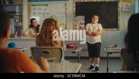 Primary School Boy Showcasing Knowledge of Ecology in Front of Class and Female Teacher. Smart Kids Sitting at the Desks During Environmental Science Lesson in Classroom. STEM Education. Static Shot. Stock Photo
