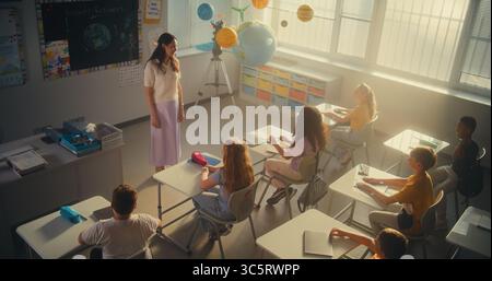Female Teacher Finishing Lesson, Announcing the End of School Day to Diverse Kids. Elementary School Students Standing Up, Packing Their Backpacks and Leaving Modern Classroom. Slow Motion. High Angle Stock Photo