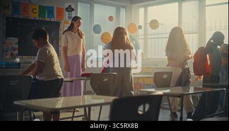 Female Teacher Finishing the Lesson, Announcing the End of School Day to Diverse Kids. Elementary School Students Standing Up, Packing Their Backpacks and Leaving Modern Classroom. Slow Motion. Stock Photo
