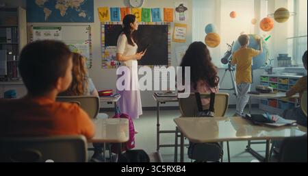 Science Lesson: Primary School Boy Presenting Renewable Energy Using Earth Planet, Talking in Front of Class. Female Teacher with Tablet Computer Educating Ecology to Smart Kids. STEM Education. Stock Photo