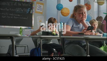 Primary School Children Eating Sandwiches, Using Smartphones and Talking with Each Other During Break. Happy Diverse Kids Sitting at Desks, Having Lunchtime Before Lesson in Modern Colorful Classroom. Stock Photo