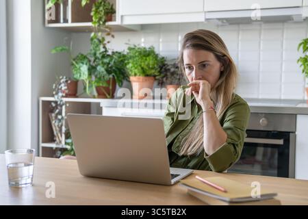 Thoughtful tired woman working on laptop, virtual classes online surrounded by books and water glass Stock Photo