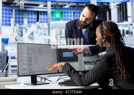 Photovoltaics factory manager coming to systems administrator desk to check progress on coding tasks. Solar panels plant team leader speaking with admin building environmentally safe solar cells Stock Photo