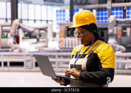 In modern facility, African american female engineer using her laptop to oversee production, ensuring efficient and safe operations. She works on creating cutting edge devices in factory. Stock Photo