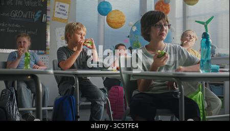 Primary School Children Sitting at the Desks, Eating Sandwiches from Lunch Boxes, Talking with Each Other During Break. Happy Diverse Kids Having Lunchtime Before Lesson in Modern Colorful Classroom. Stock Photo