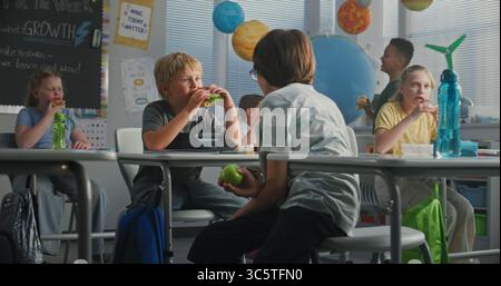 Primary School Children Sitting at the Desks, Eating Sandwiches from Lunch Boxes, Talking with Each Other During Break. Happy Diverse Kids Having Lunchtime Before Lesson in Modern Colorful Classroom. Stock Photo