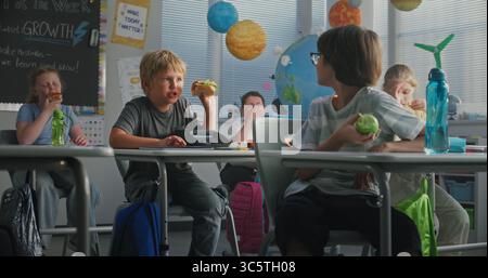 Primary School Children Sitting at the Desks, Eating Sandwiches from Lunch Boxes, Talking with Each Other During Break. Happy Diverse Kids Having Lunchtime Before Lesson in Modern Colorful Classroom. Stock Photo