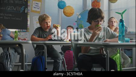 Primary School Children Sitting at the Desks, Eating Sandwiches from Lunch Boxes, Talking with Each Other During Break. Happy Diverse Kids Having Lunchtime Before Lesson in Modern Colorful Classroom. Stock Photo
