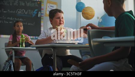 Primary School Boy Eating Sandwich from Lunch Box, Talking to Classmate and Sharing Cookie with Him. Happy Diverse Kids Having Lunchtime Sitting at the Desks in Multiethnic Classroom During Break. Stock Photo