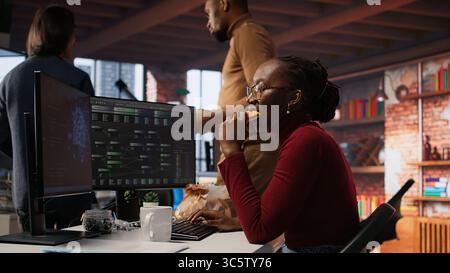 Programmer coding AI algorithm in startup, eating food at desk while refining machine learning models. Stressed woman using coding platform in Silicon Valley office and enjoying snack, camera A Stock Photo
