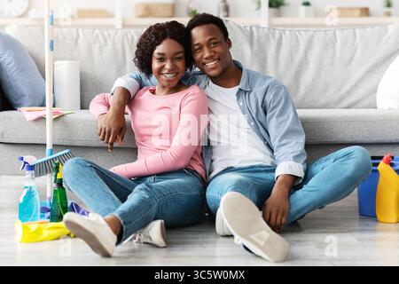 Tired black couple sitting on floor with cleaning tools Stock Photo