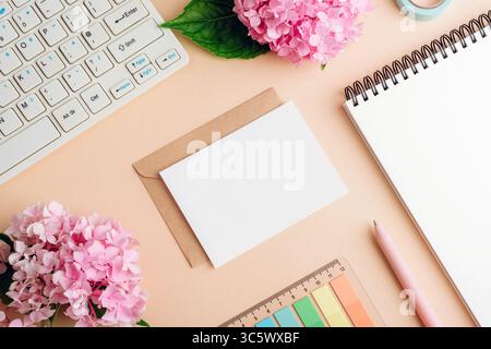Home office desktop with computer keyboard, notepad, envelope with blank card, pen, color palette and hydrangea pink flowers on beige background. Top Stock Photo