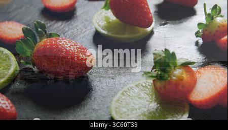 Displaying ripe red strawberry perched on lime slices on dark slate board, with sliced strawberries Stock Photo