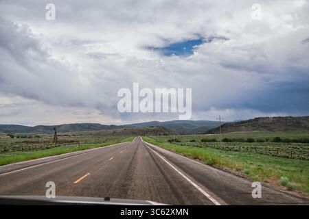 A straight two-lane highway stretches into the distance beneath a dramatic sky filled with layered clouds. Lush green pastures flank both sides of the Stock Photo