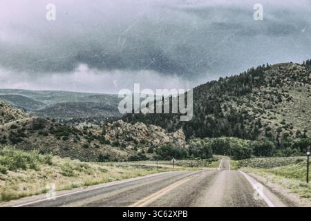 A winding two-lane road cuts through rugged hills and forested slopes under a dark, moody sky. This scenic mountain landscape in the American West evo Stock Photo