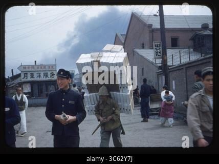 A Korean laborer ('papasan') carrying an enormous load on an A-frame back carrier through a busy street in Inchon, South Korea, as a student in uniform and other civilians pass by. May 24, 1955. Stock Photo