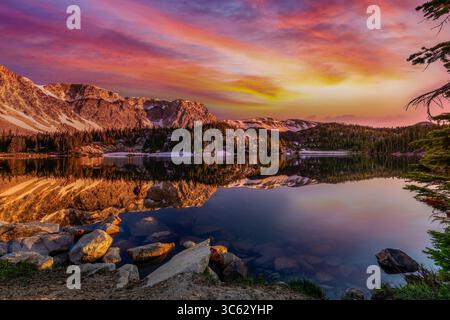 As the sun sets behind the Snowy Range, Mirror Lake in Wyoming transforms into a canvas of vibrant color. The fiery sky reflects across the calm alpin Stock Photo