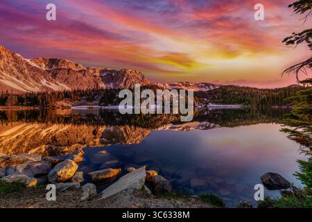 As the sun sets behind the Snowy Range, Mirror Lake in Wyoming transforms into a canvas of vibrant color. The fiery sky reflects across the calm alpin Stock Photo