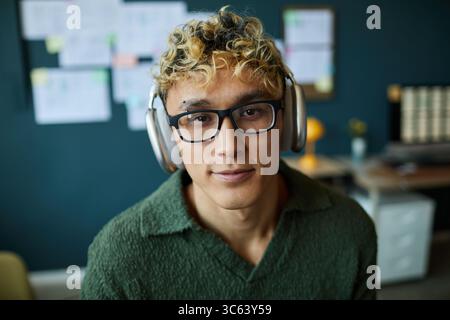 Portrait of young adult Caucasian man with curly blond hair and glasses, wearing headphones looking into camera sitting in modern office environment with blurred background Stock Photo