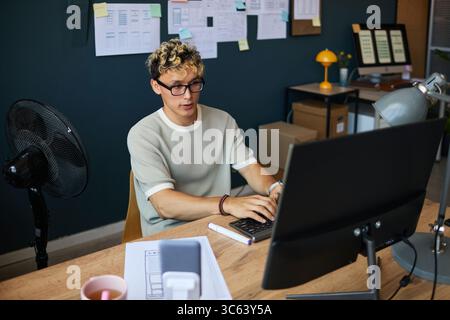 Young adult Caucasian man wearing glasses working at desk using desktop computer in modern office setting, typing on keyboard with focused expression, documents and fan visible Stock Photo