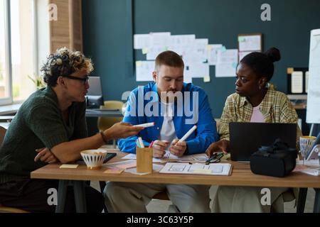 Multiethnic group of young adult colleagues collaborating at desk, discussing project ideas with documents and laptop, Caucasian man listening to Black woman explaining concept Stock Photo