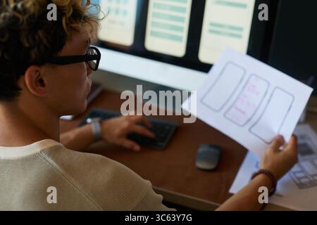 Young adult Caucasian man sitting at desk working on user interface design, holding paper with smartphone wireframes while looking at computer monitor displaying digital layouts Stock Photo