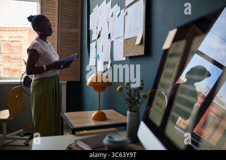 Black young adult woman standing in modern office holding documents, analyzing notes and charts pinned on wall, working on project planning and organization, computer monitor in foreground Stock Photo