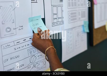 Black woman hand holding sticky note with word size written on it, placing note on wall covered with wireframe sketches and user interface design layouts during creative planning session Stock Photo
