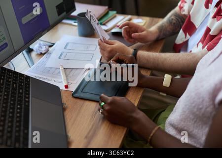 Young adult Black woman and young adult Caucasian man collaborating on digital tablet and reviewing wireframe sketches at desk, working together on user interface design project Stock Photo