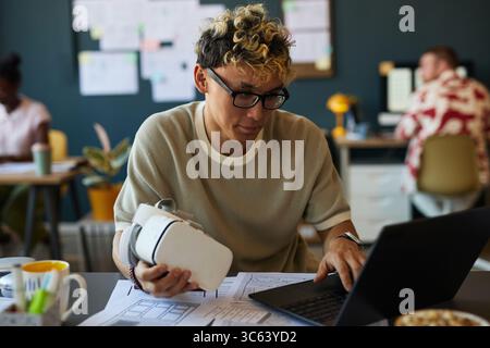 Young adult Caucasian man wearing glasses working on laptop while holding virtual reality headset in modern office setting, focusing on digital project with documents on desk Stock Photo