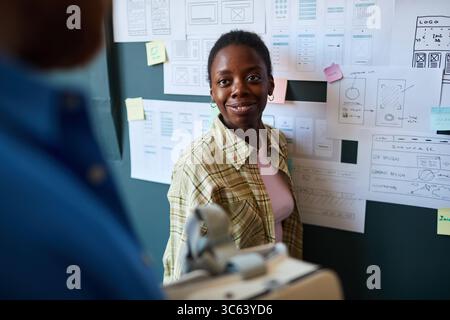 Young Black woman standing in front of wireframe sketches on wall, smiling and interacting with colleague, engaging in collaborative design process in creative workspace Stock Photo
