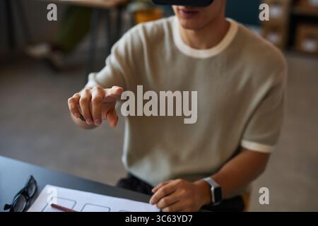 Young adult man interacting with virtual interface using hand gestures while wearing virtual reality headset, sitting at desk with smartwatch visible, engaging with digital technology Stock Photo