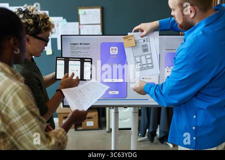Multiethnic group of young adult men and women collaborating on mobile app interface design, reviewing wireframes and digital tablet in modern office setting, standing near large display Stock Photo