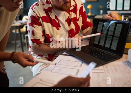 Young adult Caucasian man and young adult Asian woman collaborating on user interface design, discussing wireframes displayed on laptop screen, holding markers and reviewing sketches Stock Photo