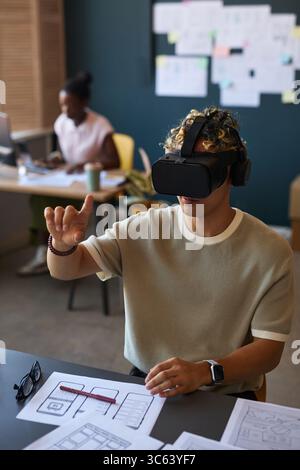 Young adult Asian man wearing virtual reality headset interacting with digital interface while sitting at desk with architectural blueprints in modern office workspace Stock Photo