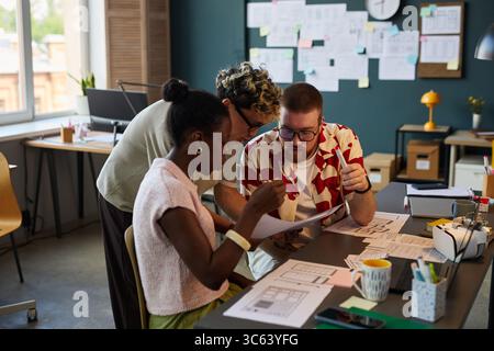 Multiethnic group of young adult colleagues collaborating on project, reviewing documents and discussing ideas at modern office desk with papers and digital devices visible Stock Photo