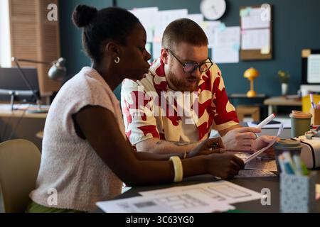 Black woman and Caucasian man collaborating at desk, reviewing documents and discussing project details in modern office setting, both focused on paperwork and teamwork Stock Photo