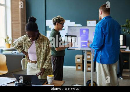 Young Black woman working on laptop while multiethnic group of young adults collaborating on digital project near interactive display in modern office workspace Stock Photo