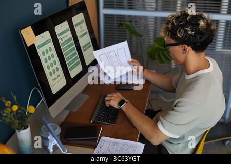 Young adult Caucasian man sitting at desk developing mobile app interface, holding wireframe sketches in one hand while working on computer with smartphone screen designs displayed Stock Photo