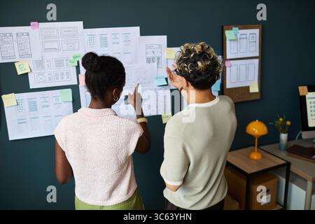 Young adult Black woman and young adult Caucasian man standing in front of wall covered with wireframes and sketches, discussing and pointing at user interface design concepts Stock Photo