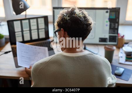 Young adult Caucasian man sitting at desk, reviewing wireframe sketches while working on computer with multiple monitors displaying user interface designs in modern office setting Stock Photo