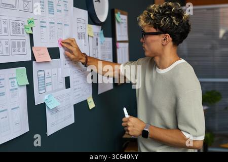 Young adult Caucasian man standing at wall, organizing paper wireframes and sticky notes, holding marker, developing user interface design, focusing on creative planning process Stock Photo