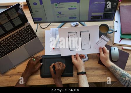 Caucasian man and Black woman collaborating on user interface design, reviewing mobile app wireframes and digital sketches at workstation with laptops and monitors displaying project details Stock Photo