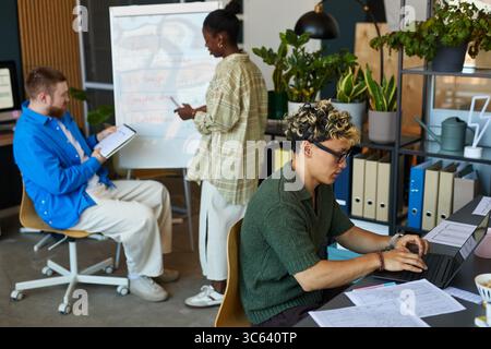 Young adult Asian man typing on laptop at desk, Black young adult woman writing on flip chart, Caucasian young adult man sitting and taking notes, diverse team collaborating in modern office Stock Photo