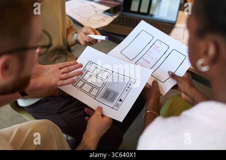 Group of young adult multiethnic men and women collaborating on mobile app interface design, holding wireframe sketches and discussing project details around table in office Stock Photo